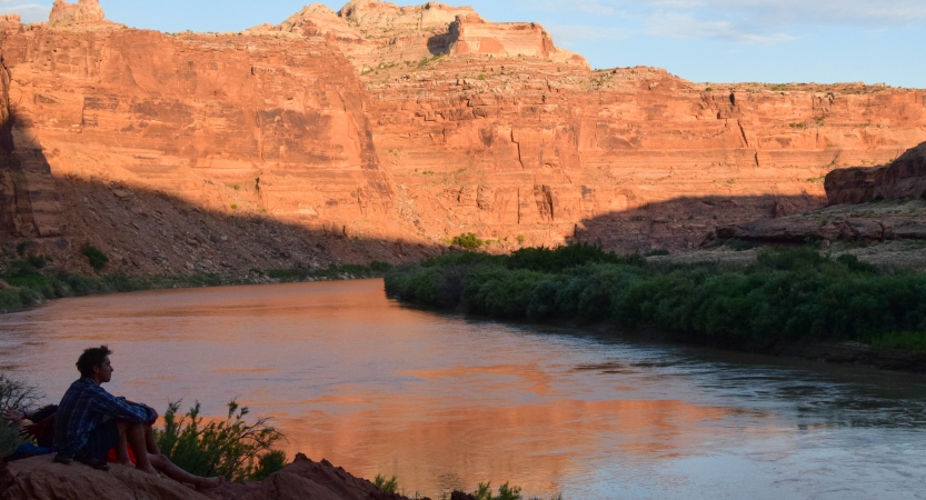 A person sits on the bank of a river, looking out over the water. In the background, there are tall red canyon walls.